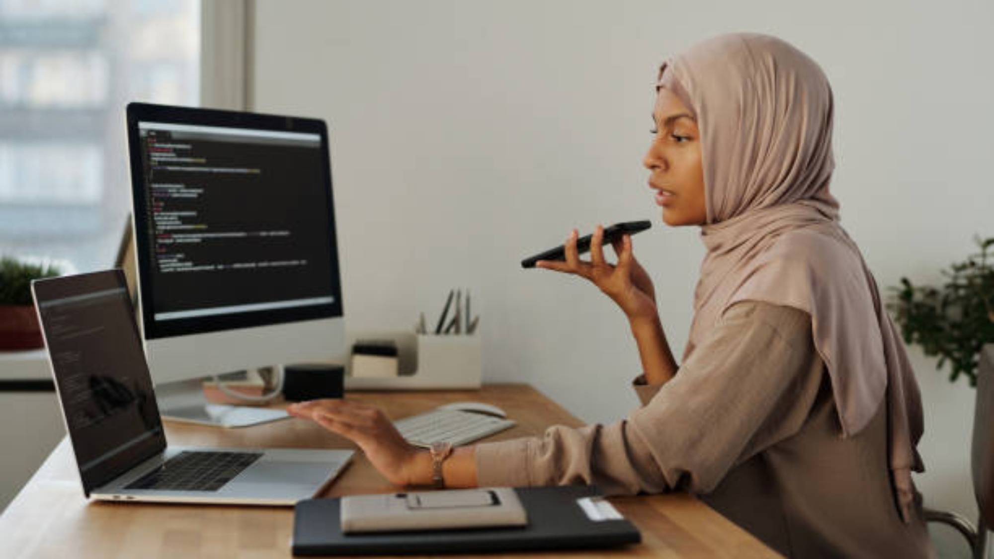 Side view of confident Muslim female programmer talking by speakerphone during work while holding mobile phone by her mouth