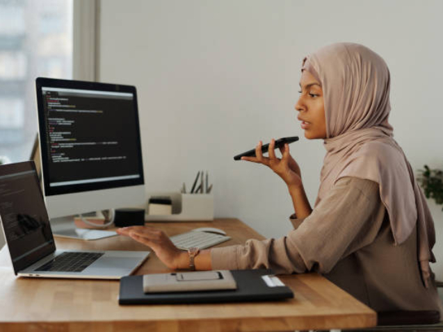 Side view of confident Muslim female programmer talking by speakerphone during work while holding mobile phone by her mouth