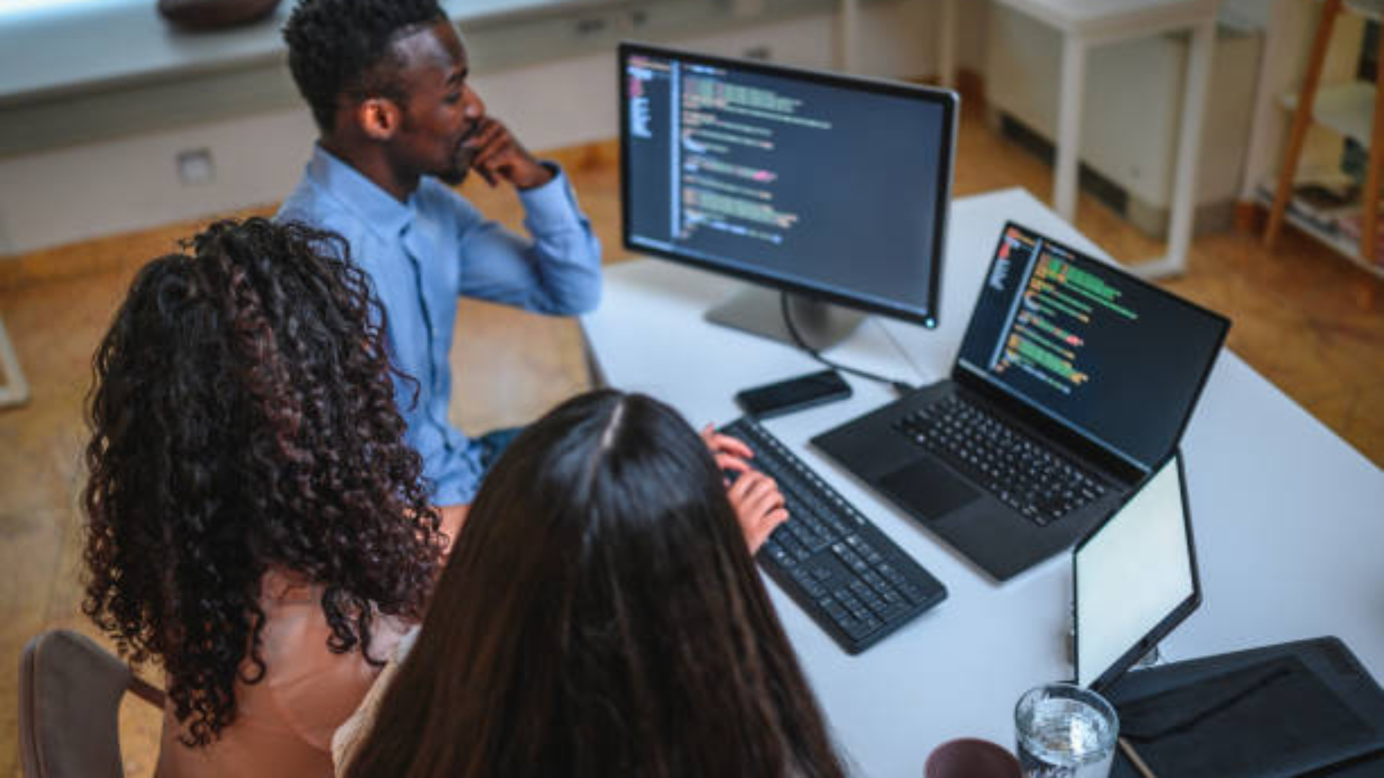 Black businessman, Asian businesswoman and Hispanic businesswoman are programming in an open plan office together. They are using computer, laptop and digital tablet.