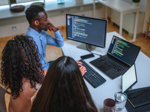 Black businessman, Asian businesswoman and Hispanic businesswoman are programming in an open plan office together. They are using computer, laptop and digital tablet.