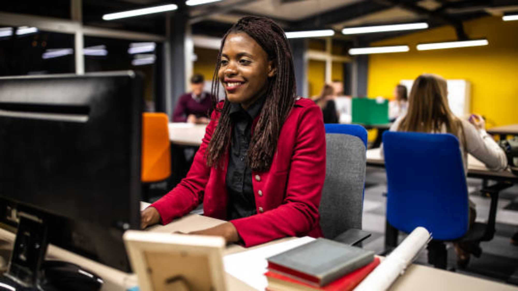 Young cheerful African woman using computer in the office, smiling and looking at papers on desk
