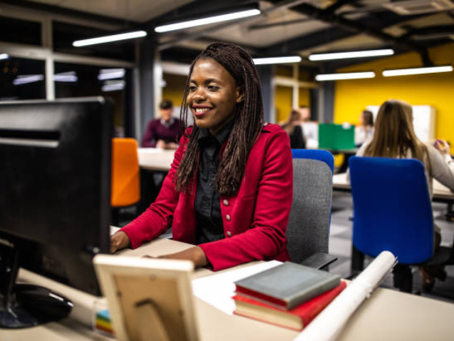 Young cheerful African woman using computer in the office, smiling and looking at papers on desk